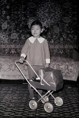 Vintage black-and-white photo of a young child in a vintage stroller with decorative luggage. The child wears a collared swea...