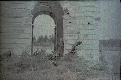 Vintage sepia-toned archway ruin with two figures standing in foreground. Stone arch, weathered brick walls, and overgrown ve...