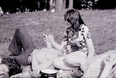 Black-and-white candid shot of a 1970s outdoor gathering: a woman in a floral blouse and patterned skirt kneels beside a recl...