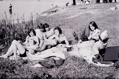 Black-and-white photo of five women lounging on a grassy lakeside picnic blanket, mid-20th century. One woman sits upright re...