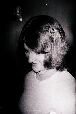 Portrait of a woman with short, voluminous bangs and a half-smile, captured in mid-conversation. She wears a high-necked, sle...