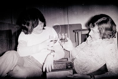 Two women share a lighthearted moment in a mid-20th-century interior, likely the 1960s–70s. One woman feeds a small dog while...