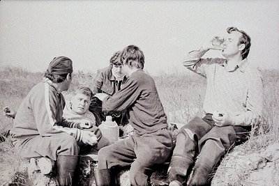 Four individuals in outdoor setting, mid-20th century. Group seated on grassy terrain, one feeding baby while others observe....