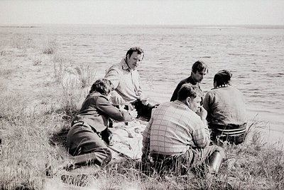 Group picnic by a lake in mid-20th century attire, likely 1950s–1960s. Five men seated on grass beside a picnic blanket, enga...