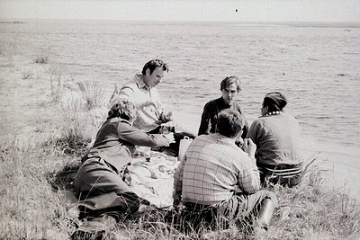 Four individuals seated outdoors by a body of water, sharing a picnic. Mid-20th century clothing suggests or . Grassland and ...