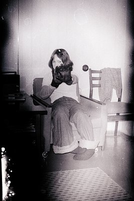 Vintage black-and-white photo of a child sitting on a wooden chair, holding a small dog. The child wears a short-sleeve shirt...