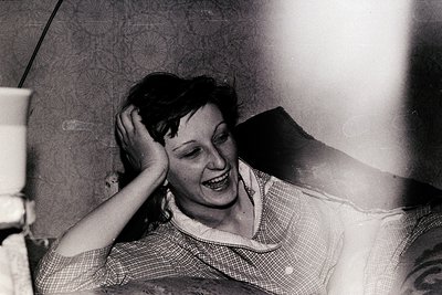 Black-and-white candid shot of a woman laughing, hand resting on her head, mid-1960s–70s. Indoor setting with patterned wallp...