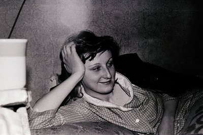 Black-and-white portrait of a woman resting her hand on her head, seated indoors. She wears a checkered blouse with a collar,...