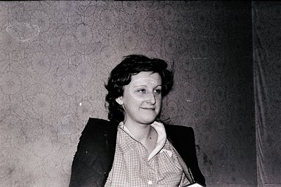Portrait of a woman in mid-20th-century fashion, wearing a checkered blouse and dark blazer. Background features patterned wa...