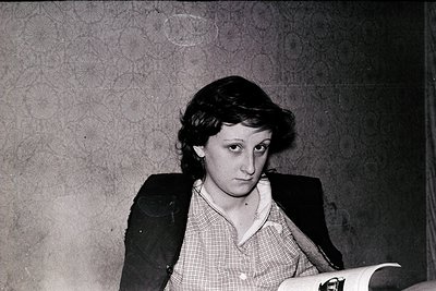 Portrait of a young woman in a mid-century indoor setting, likely 1950s–1960s. She wears a dark blazer over a checkered blous...