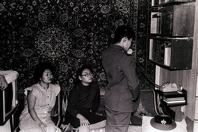 Vintage black-and-white interior shot of three individuals in a mid-century living space. Woman in floral dress sits on a pad...