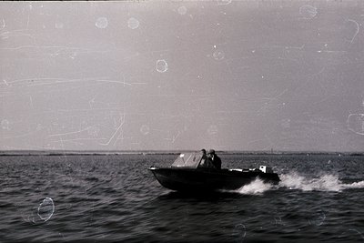 Vintage black-and-white photo of a small motorboat cutting through choppy waters, with a lone figure seated at the helm. Worn...