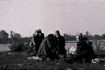 Vintage black-and-white photo of four individuals seated outdoors near a body of water, likely a lake or river. They appear t...