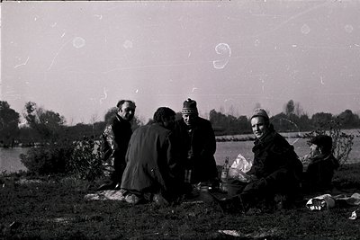 Vintage black-and-white photo of five individuals seated outdoors by a body of water, likely a lake or river. Mid-20th centur...