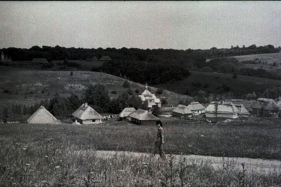 Traditional thatched-roof huts clustered on rolling hills, surrounded by dense forest. Likely Eastern European rural village,...