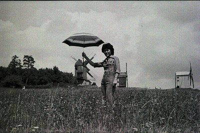 A woman in a striped blouse and overalls poses in an open field with a vintage windmill and industrial structures. Black-and-...