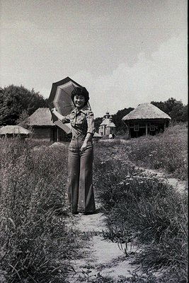 Black-and-white portrait of a woman in 1970s-style wide-leg trousers and a short-sleeve blouse, holding a large, open parasol...