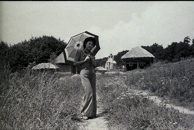 Black-and-white portrait of a woman in a traditional long-sleeved dress holding a patterned parasol, standing on a grassy hil...