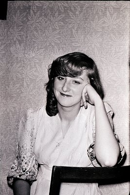Portrait of a woman in a floral lace blouse, leaning on a wooden chair against a patterned wallpaper backdrop. Mid-20th centu...