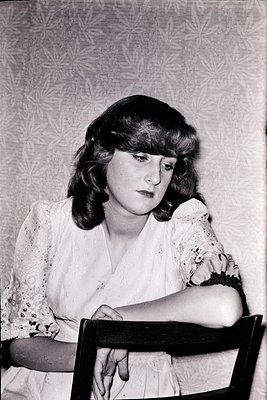 Portrait of a woman in a 1970s-style blouse with lace detailing, seated on a wooden chair. Her short, layered hair and makeup...