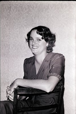 Portrait of a woman in a vintage 1940s-50s dress with a V-neck and ruffled cuffs, seated on a wooden chair against a patterne...
