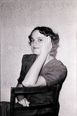 Vintage portrait of a woman in 1930s–1940s fashion, seated in a simple wooden chair against a patterned wall. She wears a fit...