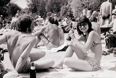 Black-and-white beach scene from the 1970s, featuring relaxed beachgoers in patterned swimwear. A woman in sunglasses and a f...