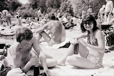 Vintage black-and-white beach scene with sunbathers lounging on towels. A man in shorts and a woman in a floral swimsuit pose...