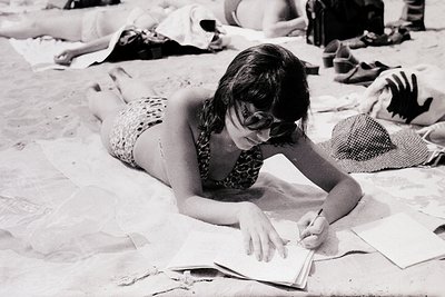 Black-and-white beach scene from the 1970s, featuring a woman writing in a notebook while lying on a towel. Patterned swimsui...