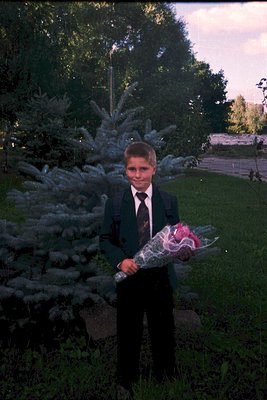 Young boy in formal attire—dark suit, white shirt, and tie—poses outdoors holding a bouquet of red and white flowers. The vin...