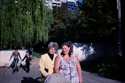 Two women in 1970s-style dresses pose near a park path with urban high-rises in background. One wears a floral sleeveless dre...