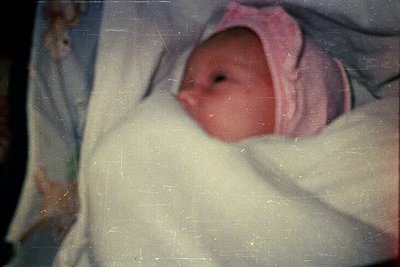 Vintage close-up of a newborn wrapped in a light blanket with a pink headband, likely mid-20th century. Soft focus and slight...