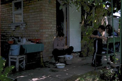 Vintage outdoor scene featuring a woman seated on a stool, washing clothes in a basin near a brick house. Two children stand ...