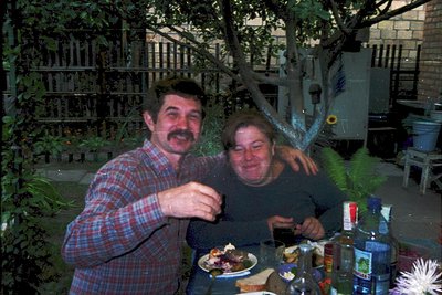 Two individuals pose indoors at a rustic wooden table, surrounded by vintage bottles and plates. The man wears a plaid shirt,...