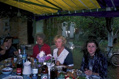 Vintage group portrait under a yellow-tinted canopy, likely 1970s–1980s. Four women seated at a table laden with bottles (inc...