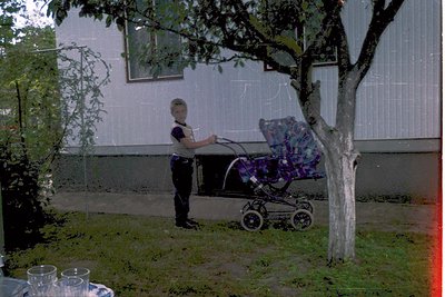 Vintage black-and-white photo of a child in 1970s-era clothing pushing a floral-patterned stroller in a residential backyard....