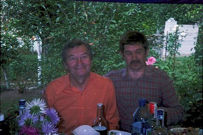 Two men pose outdoors at a garden table, surrounded by vintage bottles and floral arrangements. The man on the left wears a b...