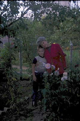 Vintage black-and-white photo of an adult and child in a lush garden, surrounded by tall plants and blooming flowers. The wom...