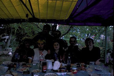 Vintage group photo under a yellow-tinted canopy, likely 1970s-80s. Six individuals pose around a table laden with food (meat...