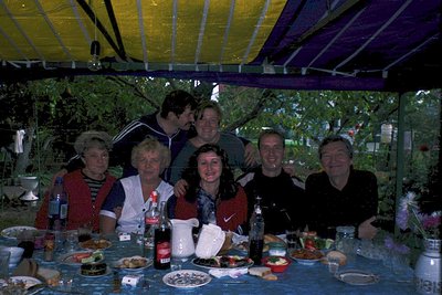 Family gathering under a striped canopy, likely 1990s–2000s. Six adults pose at a table set with bottled drinks, plates of fo...