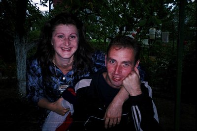 Two individuals pose indoors under low lighting, likely late 1990s–early 2000s. Woman wears a tie-dye shirt with a graphic pr...