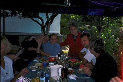 Group of six adults gathered around a long table at an outdoor dining setting, likely a garden or patio. The table is set wit...