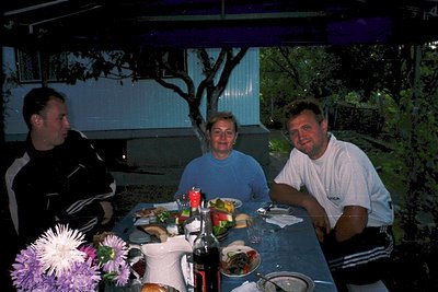 Three adults seated at an outdoor table, likely mid-1990s to early 2000s. Table set with plates, cutlery, and a bottle of win...