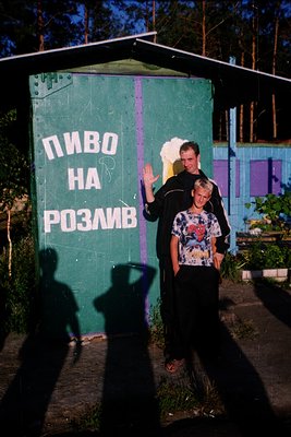 Two men pose in front of a green metal wall advertising "пиво на розлив" (beer on tap) in Cyrillic script. The man on the lef...