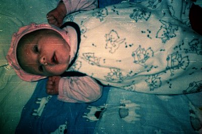 Vintage photo of an infant in a pink knit hat and blue onesie with cartoon characters, lying on a patterned blanket. Soft foc...