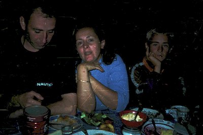Vintage black-and-white photo of three seated individuals at a table, likely from the 1970s. Central figure wears a blue shir...