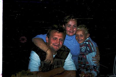 Vintage family portrait with polaroid-style grain, likely 1970s–1980s. Three individuals pose indoors, arms wrapped in a warm...