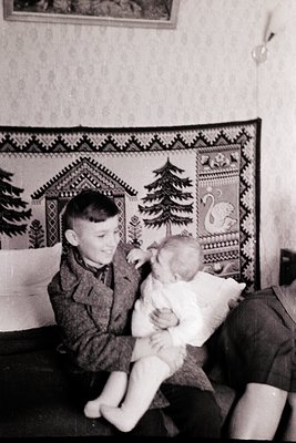 A young boy in 1950s-era clothing cradles an infant in a cozy indoor setting. Decorative wall tapestry features geometric pat...