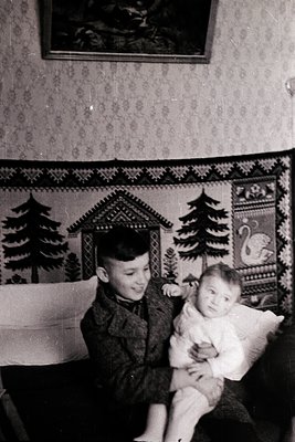 Vintage black-and-white photo of a young boy holding an infant in a cozy indoor setting, likely mid-20th century. Patterned w...