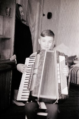 Vintage black-and-white photo of a young boy holding a large accordion, likely mid-20th century. Indoor setting with visible ...